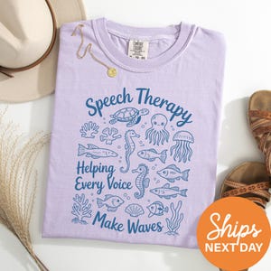 a purple t-shirt with a graphic design of marine life and the text "Speech Therapy Helping Every Voice Make Waves" printed on it. The t-shirt is laid out on a white surface, along with a beige hat, a pair of sandals, and some plants.