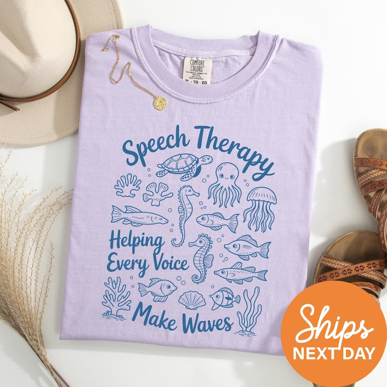 a purple t-shirt with a graphic design of marine life and the text "Speech Therapy Helping Every Voice Make Waves" printed on it. The t-shirt is laid out on a white surface, along with a beige hat, a pair of sandals, and some plants.