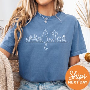 A woman wearing a blue t-shirt with a white cat silhouette and various potted plants printed on it.