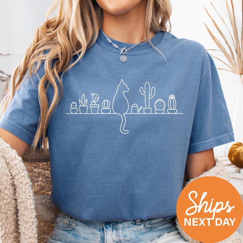 A woman wearing a blue t-shirt with a white cat silhouette and various potted plants printed on it.