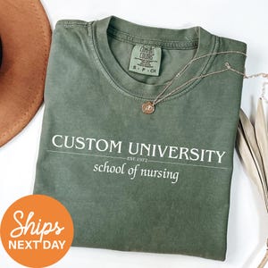May include: An olive green t-shirt with the words "CUSTOM UNIVERSITY" and "school of nursing" printed in white. A gold necklace with a pendant rests on the shirt. The shirt is folded, and a brown hat is partially visible.