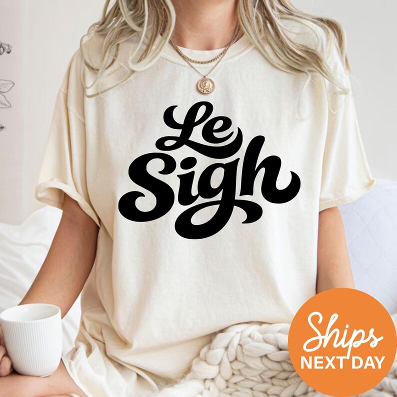 A person wearing a white t-shirt with the text "Le Sigh" printed on it, sitting on a bed with a white blanket.