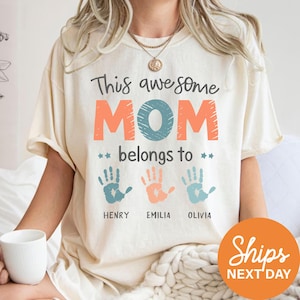 A person wearing a white t-shirt with a graphic design that says "This awesome MOM belongs to HENRY, EMILIA, OLIVIA" along with handprints.
