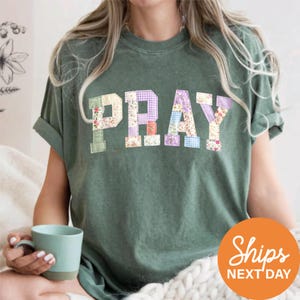 May include: Green t-shirt with the word "PRAY" in patchwork letters. The letters are made of various floral and patterned fabrics. A light blue mug is held in the hand. The shirt has rolled-up sleeves.