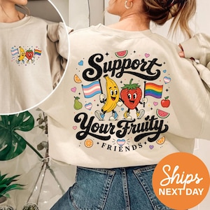 May include: A beige crewneck sweatshirt featuring a large graphic on the back with a cartoon banana and strawberry holding pride flags. The text reads Support Your Fruity Friends, surrounded by small fruit and heart icons. A smaller version is on the front.