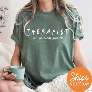 A woman wearing a green t-shirt with the text "THERAPIST I'LL BE THERE FOR YOU" printed on it, sitting on a couch with a cup of coffee in her hand.