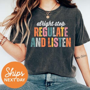May include: Dark gray t-shirt with the words "alright stop REGULATE AND LISTEN" in colorful block letters. The shirt has a crew neck and short sleeves. The text is in white, orange, yellow, pink, teal, and blue.