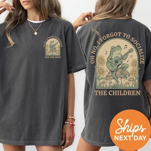 a woman wearing a gray t-shirt with a graphic design on the back. The design includes a frog playing a guitar and the text "OH NO, I FORGOTT TO SOCIALIZE THE CHILDREN".