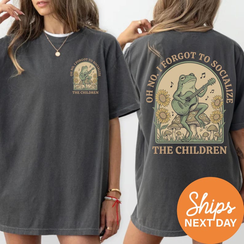 a woman wearing a gray t-shirt with a graphic design on the back. The design includes a frog playing a guitar and the text "OH NO, I FORGOTT TO SOCIALIZE THE CHILDREN".