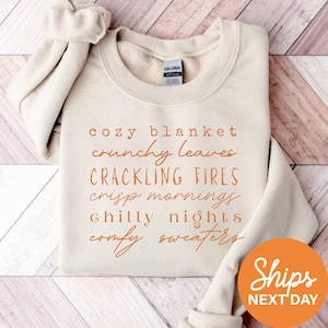 May include: A cream-colored crewneck sweatshirt featuring orange text that reads: cozy blanket, crunchy leaves, CRACKLING FIRES, crisp mornings, chilly nights, comfy sweaters. The garment has a Gildan brand label inside the collar.