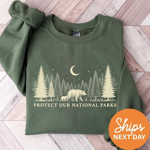 May include: Green sweatshirt with a forest scene featuring a bear and cub, a crescent moon, and the text "PROTECT OUR NATIONAL PARKS". The design is in white. The sweatshirt has long sleeves.