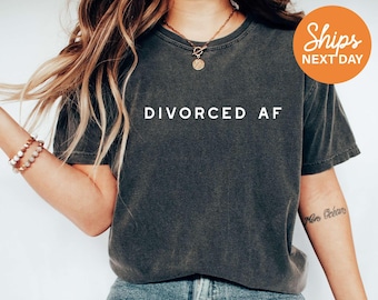 May include: A charcoal grey short-sleeved t-shirt featuring the text DIVORCED AF printed in white capital letters across the chest. The garment has a crew neckline and a washed, vintage-style finish, paired with blue denim jeans.