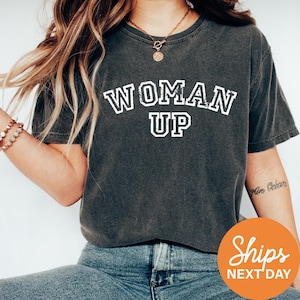 A woman wearing a gray t-shirt with the text "WOMAN UP" printed on it, along with a necklace and bracelets.