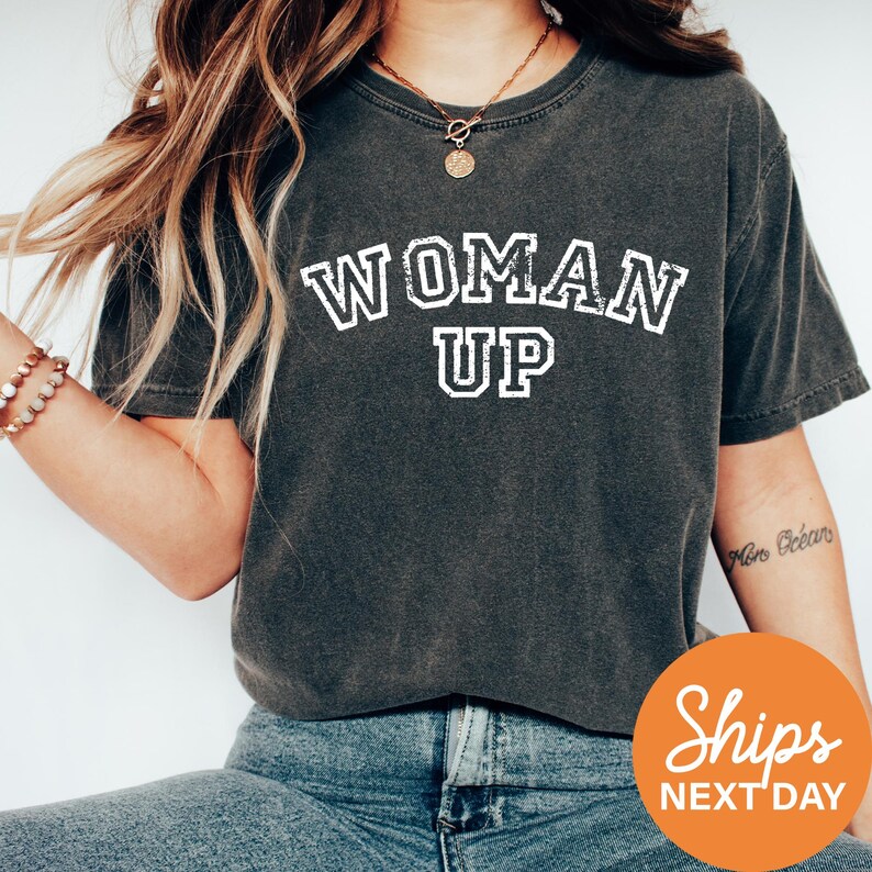 A woman wearing a gray t-shirt with the text "WOMAN UP" printed on it, along with a necklace and bracelets.