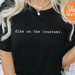 May include: A black short-sleeved t-shirt featuring the white text "dibs on the (custom)." printed across the chest. The garment has a classic crew neckline and a simple, solid-colored design, providing a clean look for casual everyday wear.