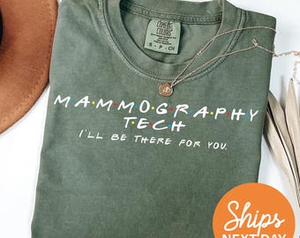 May include: An olive green short-sleeve t-shirt with white text that reads MAMMOGRAPHY TECH and I'LL BE THERE FOR YOU. The text features small colored dots between the letters. A gold-toned necklace with a circular pendant rests on the shirt.