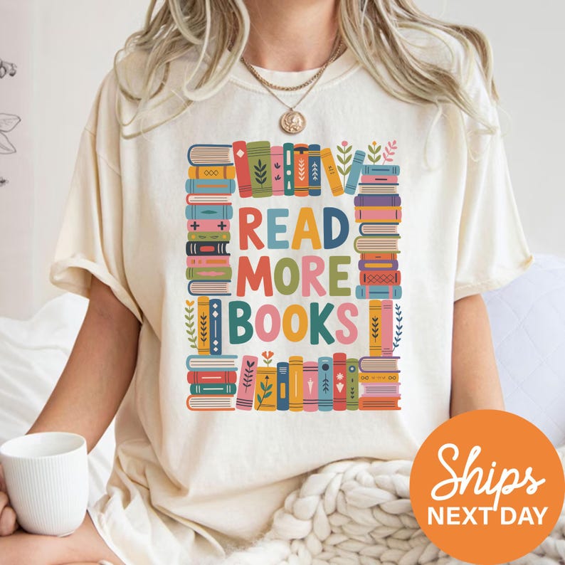 A person wearing a white t-shirt with a colorful graphic design that says "READ MORE BOOKS" surrounded by various books and illustrations.