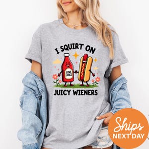 A woman wearing a gray t-shirt with a graphic design featuring two cartoon hot dogs, one red and one yellow, along with the text "I SQUIRT ON JUICY WIENERS".