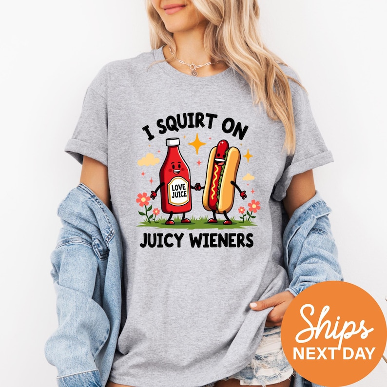 A woman wearing a gray t-shirt with a graphic design featuring two cartoon hot dogs, one red and one yellow, along with the text "I SQUIRT ON JUICY WIENERS".