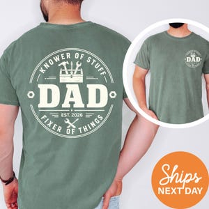 May include: A sage green short-sleeve t-shirt featuring a large white circular graphic on the back with a toolbox, tools, and the text "KNOWER OF STUFF DAD FIXER OF THINGS EST. 2026". A smaller version of the same graphic is printed on the front left chest.
