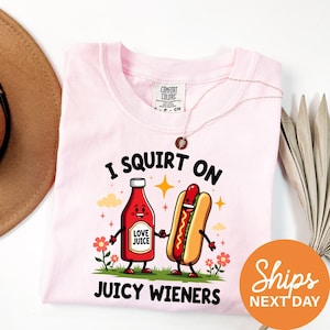 a pink t-shirt with a cartoon illustration of a hot dog and a bottle of ketchup, along with the text "I SQUIRT ON JUICY WIENERS" and a tagline "Shops NEXT DAY".