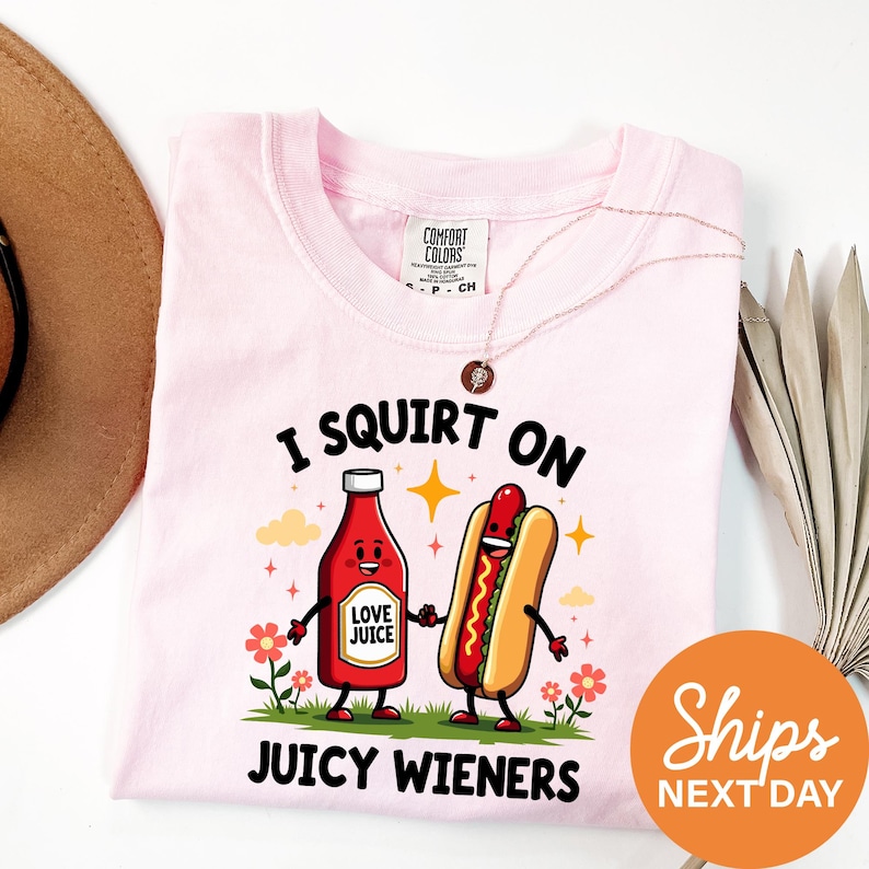 a pink t-shirt with a cartoon illustration of a hot dog and a bottle of ketchup, along with the text "I SQUIRT ON JUICY WIENERS" and a tagline "Shops NEXT DAY".