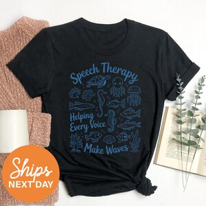 a black t-shirt with a graphic design of marine life and the text "Speech Therapy Helping Every Voice Make Waves" printed on it. The t-shirt is placed on a white surface, along with a beige sweater, a book, and some greenery.