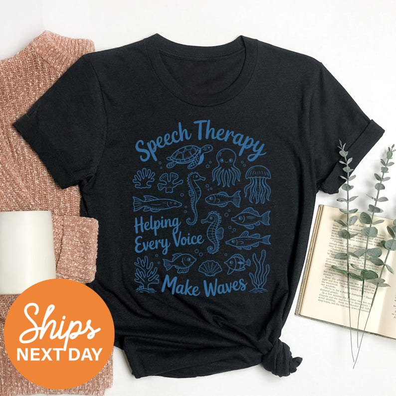 a black t-shirt with a graphic design of marine life and the text "Speech Therapy Helping Every Voice Make Waves" printed on it. The t-shirt is placed on a white surface, along with a beige sweater, a book, and some greenery.