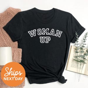 a black t-shirt with the text "WOMAN UP" printed on it, along with a sweater and a book.