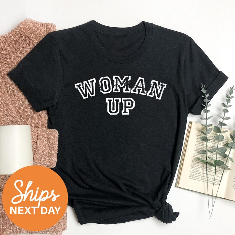 a black t-shirt with the text "WOMAN UP" printed on it, along with a sweater and a book.
