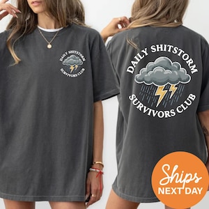 a woman wearing a gray t-shirt with a graphic design on the back. The design includes a cloud with lightning bolts, representing the "Daily Shitstorm Survivors Club".