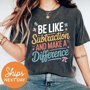 May include: Dark gray t-shirt with the text "BE LIKE Subtraction AND MAKE A Difference" in white, yellow, blue, and pink. The shirt has a crew neck and short sleeves. The shirt is made of a soft material.