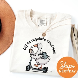May include: Cream-colored t-shirt with a graphic of a goose riding a scooter, holding a coffee cup, and wearing a backpack. The text "Off to regulate emotions" is printed around the goose. A gold necklace and a brown hat are visible.