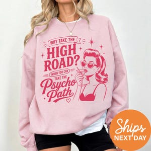 May include: A light pink crewneck sweatshirt featuring a dark pink graphic print of a person wearing sunglasses and holding a martini glass. The text reads: Why take the high road? When you can take the psycho path. The design includes star accents.