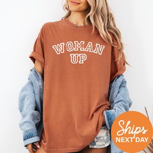 A woman wearing a brown t-shirt with the text "WOMAN UP" printed on it, along with a denim jacket, stands against a white background.
