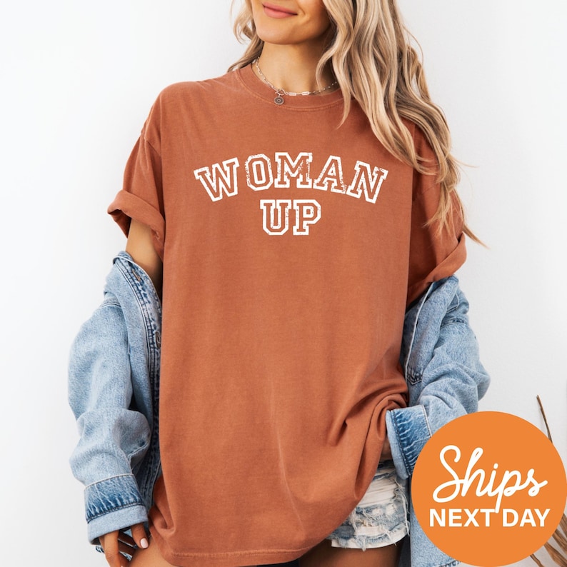 A woman wearing a brown t-shirt with the text "WOMAN UP" printed on it, along with a denim jacket, stands against a white background.