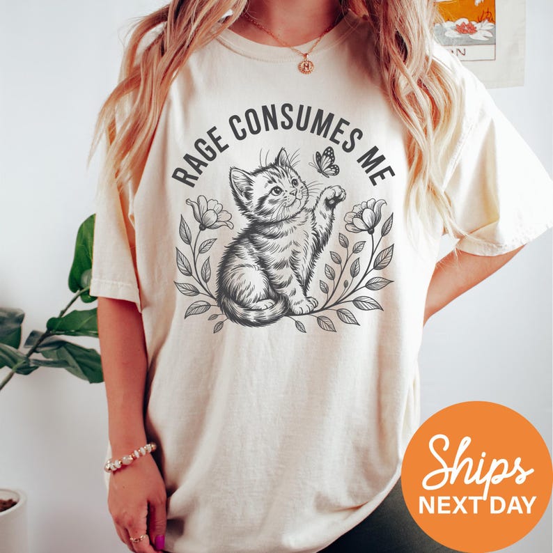 A person wearing a white t-shirt with a graphic design of a cat and flowers, along with the text "RAGE CONSUMES ME" printed on it.