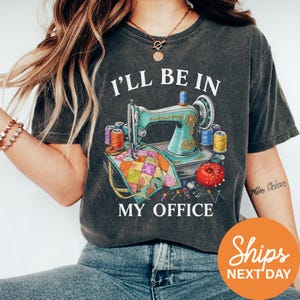A woman wearing a gray t-shirt with a graphic design of sewing equipment and the text "I'll be in my office" printed on it.