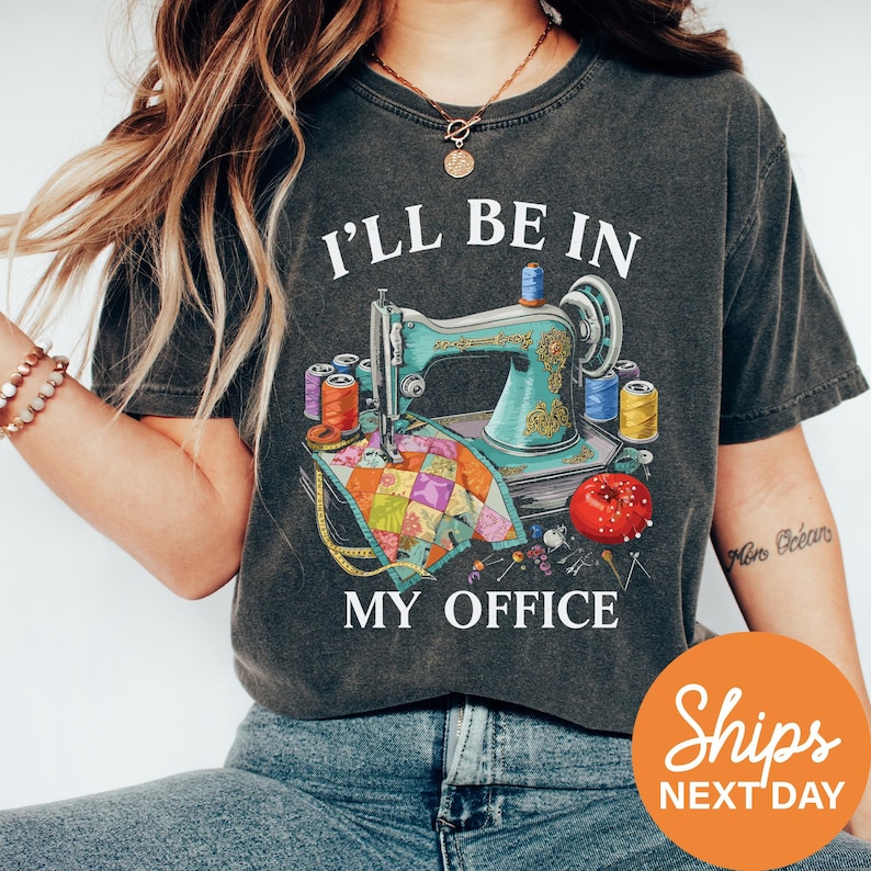 A woman wearing a gray t-shirt with a graphic design of sewing equipment and the text "I'll be in my office" printed on it.