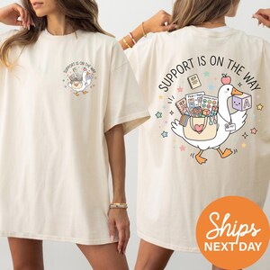 two white t-shirts with a graphic design on the back. The design includes a cartoon goose carrying a basket of items, with the text "Support is on the way" written around the goose.