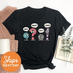 a black t-shirt with a graphic design depicting four cartoon characters with speech bubbles saying "WAIT", "WHAT?", "I AGREE!", and "STOP IT". The characters are arranged in a row, and the t-shirt is placed on a white surface alongside some other items, such as a sweater and a book.