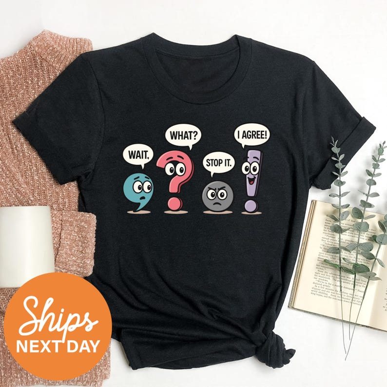 a black t-shirt with a graphic design depicting four cartoon characters with speech bubbles saying "WAIT", "WHAT?", "I AGREE!", and "STOP IT". The characters are arranged in a row, and the t-shirt is placed on a white surface alongside some other items, such as a sweater and a book.