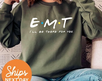 May include: An olive green crewneck sweatshirt featuring the white text E M T with a blue dot between E and M and a yellow dot between M and T. Below the text, the phrase I'LL BE THERE FOR YOU is printed in white capital letters.