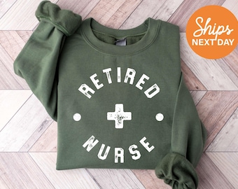 May include: An olive green crewneck sweatshirt featuring white distressed lettering that reads RETIRED NURSE in a circular arrangement, with a central white cross symbol and two small white dots on either side of the cross.