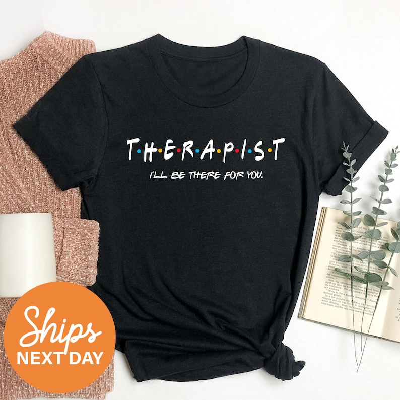 a black t-shirt with the text "THERAPIST I'LL BE THERE FOR YOU" printed on it, along with a brown sweater and a book.