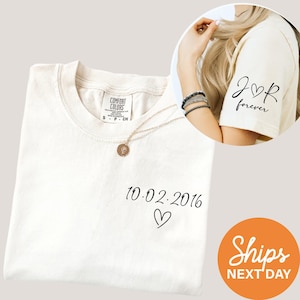 May include: Cream-colored t-shirt with the date "10.02.2016" and a heart symbol printed in black. The sleeve of the shirt features the text "J♡R forever" in black. A gold necklace with a pendant is draped on the shirt.