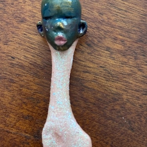 May include: An abstract figurine with a human-like face. The head is dark green with gold accents, featuring closed eyes and pink lips. The neck and base are light pink with glitter, creating a unique, artistic piece.
