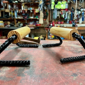 Peut inclure: Deux porte-serviettes faits à la main de style rustique. Chacun a une poignée cylindrique en bois clair fixée à une base métallique noire incurvée et texturée. Les supports sont présentés sur une surface rouge.