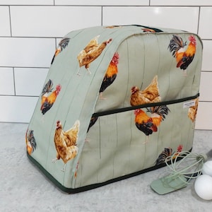 May include: A light green appliance cover with a repeating pattern of brown chickens and roosters. The cover has a dark green trim and a pocket on the front. The background has vertical lines. The cover is on a light grey surface.