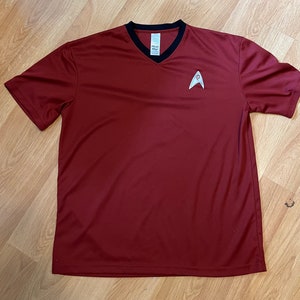 May include: A maroon short-sleeved t-shirt with a black v-neck collar and a silver Starfleet insignia on the front.